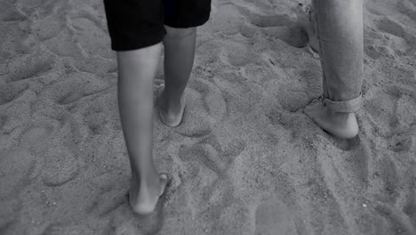 A-black-and-white-image-of-the-feet-of-a-child-with-his-mother,-walking-on-the-beach