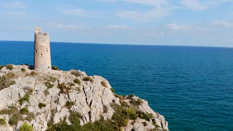 Drone-flies-over-a-coastal-watchtower,-centuries-old-tower-on-cliff-the-sea-and-blue-sky-with-rocks-in-lateral-traveling