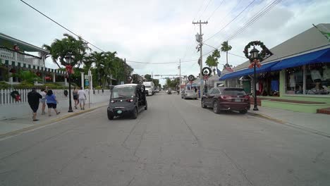 Key-West-Street-Mit-Straßenlaternen,-Die-Mit-Weihnachtskränzen-Geschmückt-Sind,-Mit-Menschen-Auf-Dem-Bürgersteig-Und-Einem-Golfwagen,-Der-Auf-Die-Kamera-Zufährt