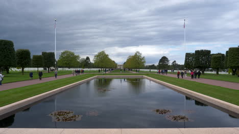 Multitud-Caminando-Alrededor-Del-Cementerio-Americano-De-Normandía-Y-La-Piscina-Reflectante-Conmemorativa,-Postes-De-Bandera-Americana-A-Ambos-Lados