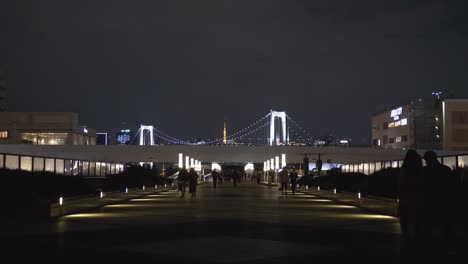 Escena-Nocturna-De-La-Pasarela-De-Odaiba-Con-Gente-Caminando-Y-El-Puente-Del-Arco-Iris-En-La-Bahía-De-Tokio-Al-Fondo,-Tokio,-Japón