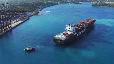 Scenic-view-of-cargo-ship-arriving-at-seaport-by-large-dockyard-cranes-being-pulled-by-tugboat,-Punta-Caucedo,-Dominican-Republic,-overhead-aerial