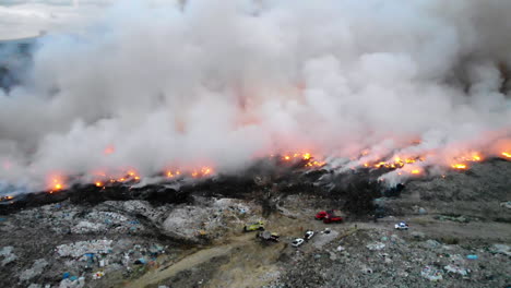 Aerial,-reverse,-drone-shot-of-a-Junkyard-on-fire,-firefighter-trucks-in-middle-of-garbage,-waste-and-trash,-toxic-blue-smoke-rising,-in-Santo-Domingo,-Dominican-Republic