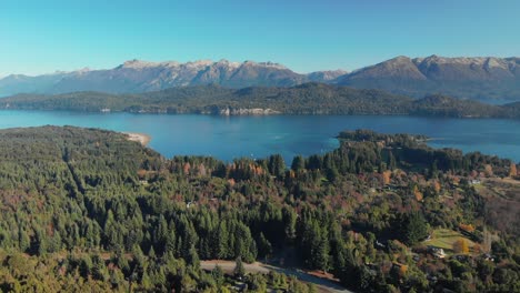 Aerial-view-of-a-forest-with-houses,-a-lake-and-the-Andes-mountains-in-the-background