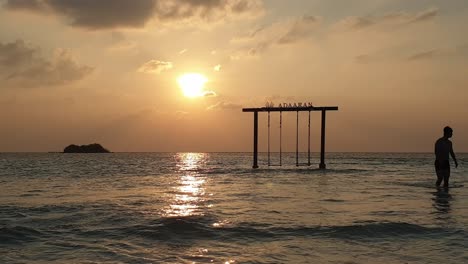 Man-walks-in-ocean-of-Maldives-with-swing-in-background-at-dusk