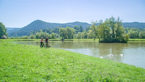 Two-caucasian-travellers-riding-bikes-alongside-a-beautiful-lake-with-swans