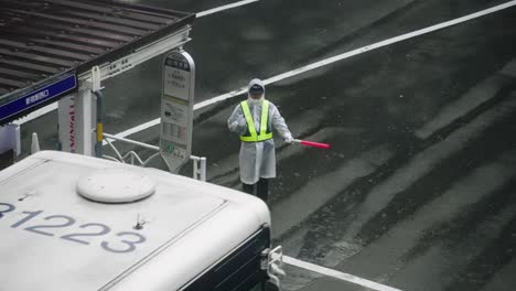 Guardia-De-Tráfico-Japonés-Usando-Impermeable-Deteniendo-Un-Autobús-En-La-Intersección-En-Tokio,-Japón-En-Temporada-De-Lluvias---ángulo-Alto,-Cámara-Lenta