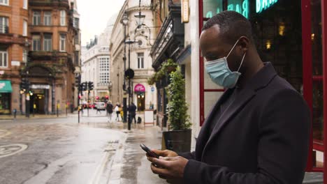 Mann,-Der-Eine-Medizinische-Maske-Trägt-Und-Ein-Smartphone-Auf-Einer-Belebten-Straße-In-London-Benutzt