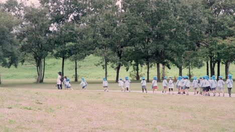 Grupo-De-Niños-De-Escuela-Primaria-Japoneses-Siguiendo-A-Su-Maestro-Y-Disfrutando-De-Su-Viaje-De-Campo-En-El-Parque-Público-De-Saitama,-Japón