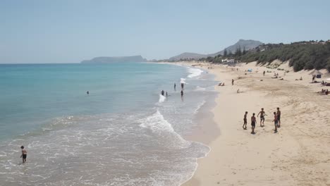 Summertime-scene-of-people-swimming,-relaxing-and-kids-playing-soccer-by-ocean-sea-waves-on-Porto-Santo-island-sandy-beach-in-Madeiran-archipelago-with-mountain-range-in-background,-Portugal,-static