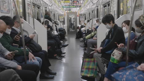 Viajeros-Locales-Con-Mascarilla-Sosteniendo-Sus-Teléfonos-Móviles-Mientras-Están-Sentados-En-Un-Tren-De-Viaje-En-Tokio,-Japón