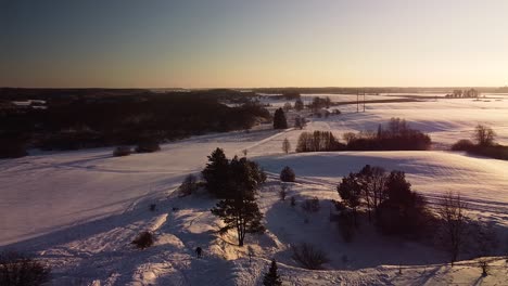 Beautiful-aerial-view-of-snow-covered-fields-near-frozen-Sventaja-river-in-sunny-winter-day,-golden-hour,-wide-angle-drone-shot-moving-bacwards