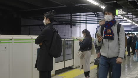 Hombres-Con-Máscara-Pasando-Pasajeros-De-Pie-En-La-Plataforma-De-La-Estación-De-Tren-En-Tokio,-Japón-Durante-La-Pandemia