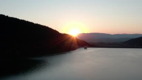 Aerial-crane-shot-of-sun-setting-behind-ridge-over-picturesque-mountain-lake