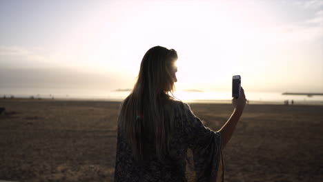 Chica-Sosteniendo-Teléfono-Tomando-Fotos-En-La-Playa-Durante-La-Puesta-De-Sol-En-Essaouira,-Marruecos