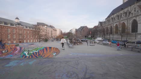 Ein-Junge-Fährt-Auf-Einem-Skateboard-Den-Skatepark-Hinunter-Und-Ein-Junger-Mann-Fährt-Einen-Roller-Im-Straßenskatepark-Neben-Der-Kapellenkirche-In-Brüssel,-Belgien