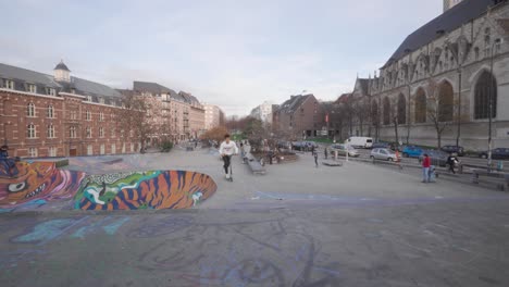 Boy-on-a-skateboard-goes-down-the-skate-park-and-a-young-man-rides-a-scooter-in-street-skate-park-next-to-the-Chapel-Church-,-Brussels,-Belgium