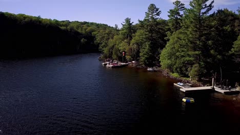 Drohne-Fliegt-über-Cottage-Docks-Am-See-Mit-Wehenden-Kanadischen-Und-Deutschen-Flaggen-Im-Wind-Und-Grünen-Kiefern-Am-Ufer