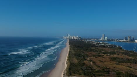 Aerial-view-of-a-coastline-near-a-large-area-of-green-space-with-a-sprawling-city-skyline-in-the-distant