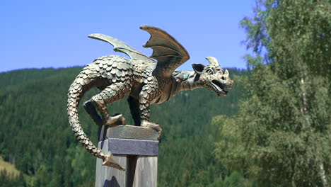 Estatua-De-Dragón-De-Bronce-En-Poste,-Paisaje-De-Montaña,-Bosque-Y-Pasto-Alpino