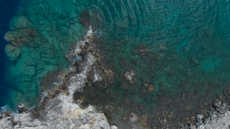 Aerial:-Drone-looking-down-to-cliffs-surrounded-by-waves-and-turquoise-sea-water