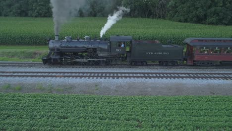 Vista-Aérea-De-Un-Motor-De-Vapor-De-1924-Con-Un-Tren-De-Pasajeros-Que-Viaja-A-Lo-Largo-De-La-Campiña-Amish-Mientras-Las-Puestas-De-Sol-En-Un-Día-De-Verano-Visto-Por-Un-Dron