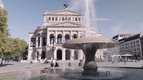 Slow-motion-of-people-walking-in-front-of-Old-Opera-house-in-Frankfurt,-Hesse,-Germany-during-daytime-with-view-of-clear-sky-and-a-water-fountain-as-foreground