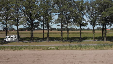 Aerial-sideways-follow-of-a-camper-passing-by-on-a-Dutch-country-road-with-trees-on-either-side-with-crop-fields-against-a-blue-sky