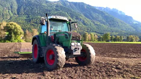 Un-Tractor-De-Cuatro-Ruedas-Con-El-Conductor-Aplanando-Tierras-Agrícolas-Con-Montañas-Y-árboles-Verdes-En-El-Fondo