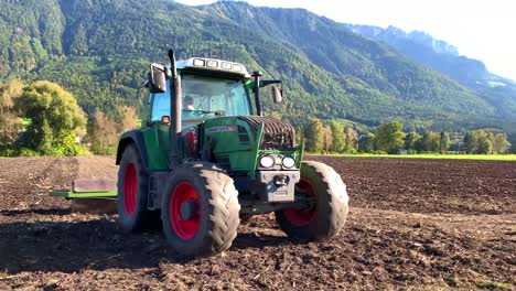 Ein-Vierradtraktor-Mit-Dem-Fahrer,-Der-Ackerland-Ebnet,-Mit-Bergen-Und-Grünen-Bäumen-Im-Hintergrund