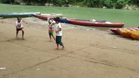 Niños-Rurales-Jugando-Cerca-Del-Río,-Barcos-Vistos-En-El-Fondo