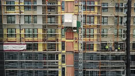 A-construction-elevator-slowly-descending-between-scaffolding-on-the-side-of-a-building---Aerial-shot