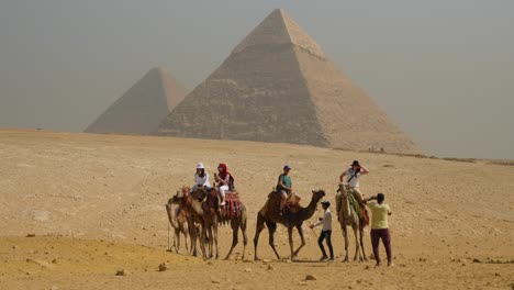 Touristen-Sitzen-Auf-Kamelen-In-Der-Wüste-Mit-Großen-Pyramiden-Im-Hintergrund