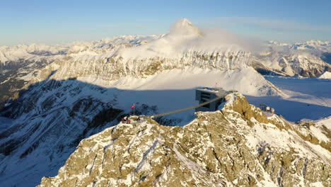 Sobrevuelo-Aéreo-Sobre-El-Mirador-Con-La-Bandera-Suiza-Ondeando-Al-Viento,-Rodeado-De-Altas-Cumbres-Alpinas-Cubiertas-De-Nieve-Y-Con-Vistas-Al-Pueblo-De-Les-Diablerets