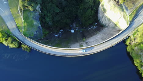 Aerial-Top-View-of-a-car-driving-across-a-Dam-with-Bluewater-and-green-forest