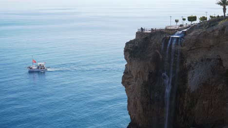 Establishing-shot,-boat-sailing-in-the-Mediterranean-sea,-waterfall-at-the-cliff-of-Antalya-Turkey