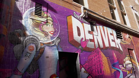 A-wall-mural-by-a-street-artist-that-says,-"greetings-from-Denver"-with-a-lady-in-an-astronaut-suit-pointing-to-it