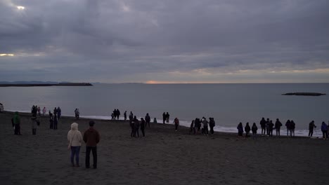 Strand-In-Japan-–-Menschen-Warten-Am-Ufer-Auf-Den-Sonnenaufgang-Mit-Plätschernden-Wellen-Und-Einer-Von-Meerwasser-Umgebenen-Insel-An-Einem-Bewölkten-Morgen-–-Weitwinkelaufnahme
