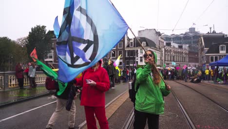 Dos-Niñas-Ondeando-Banderas-Con-Un-Grupo-De-Manifestantes-En-La-Parte-De-Atrás-En-El-Puente-Blauwbrug-Durante-La-Protesta-Climática-De-La-Rebelión-De-Extinción-En-Amsterdam,-Países-Bajos
