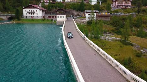 Lake-Fedaia-Dam-in-the-Dolomite-mountains-with-a-black-car-passing-the-road-above-the-structure,-Aerial-Drone-orbit-tracking-shot