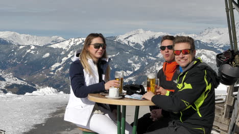 Medium-wide-shot-of-a-Caucasian-woman-and-two-Caucasian-men-enjoying-beer-at-a-mountain-ski-resort-in-Wagrain-with-beautiful-snow-covered-mountains-in-the-background