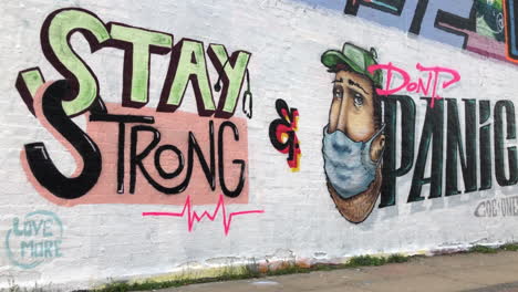 People-walk,-cycle-and-run-past-a-piece-of-graffiti-art-on-a-wall-of-a-character-wearing-a-surgical-face-mask-and-the-defiant-anti-Coronavirus-message-“stay-strong-and-don’t-panic”