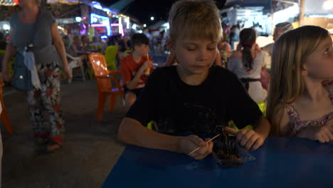 Junge-Isst-Einen-Skorpion,-Begleitet-Von-Seiner-Familie,-Auf-Einem-Lokalen-Markt-In-Khao-Lak,-Thailand