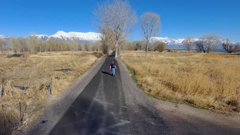 Mature,-balding-man-walking-with-a-backpack-down-a-nature-trail-with-the-snow-capped-mountains-in-the-distance---tilting-aerial-view