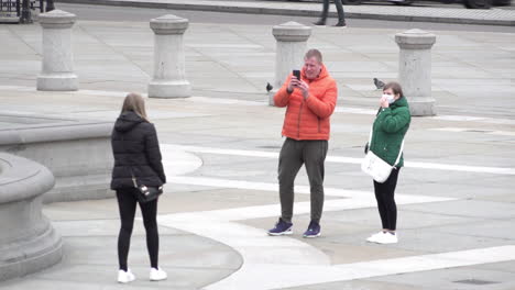 A-group-of-tourists-take-turns-to-have-their-photos-taken-in-Trafalgar-Square,-the-one-wearing-a-surgical-face-mask-removes-it-to-have-her-photo-taken