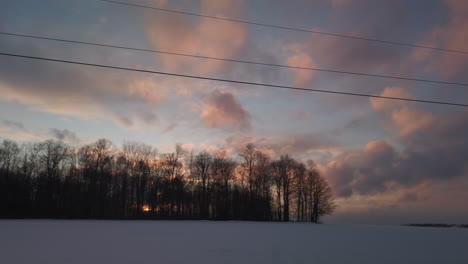 Wide-sunset-travelling-shot-of-farm-fields-in-winter,-with-big-sky-and-sun-shining-through-the-trees-in-the-forest