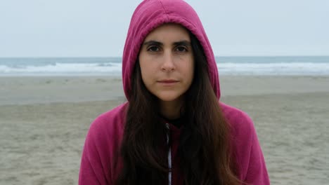 Close-up-portrait-of-casual-brunette-young-woman-smiling-with-a-hood-looking-at-camera-at-the-beach,-ocean-background