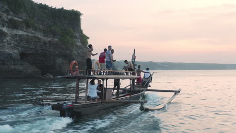 Chasing-behind-a-group-of-young-men-on-a-local-party-boat-during-sunset,-Bali,-aerial-follow