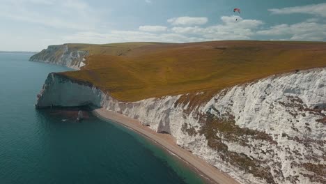Aerial-shot-of-Jurassic-Coast-and-revealing-a-paraglider-flying-over-the-white-cliffs-near-Dorset,-UK