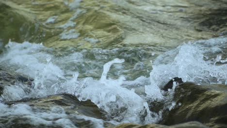 Close-up-of-wave-breaking-on-dark-rocks,-splashing-water-in-the-air,-waterdrops